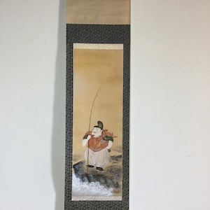 May include: A vertical hanging scroll featuring a painting of a figure in traditional attire standing on a rocky shore with a fishing rod. The figure wears a red garment and light-colored trousers. The scroll has dark patterned borders and tan ends.