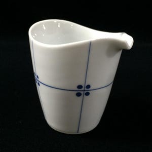 Può includere: Una lattiera in ceramica bianca caratterizzata da un delicato motivo a griglia blu con piccoli punti in ogni intersezione. Il recipiente presenta un corpo liscio e affusolato con un beccuccio pronunciato su un lato per versare liquidi.