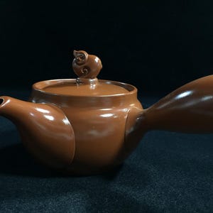 May include: A brown ceramic teapot with a side handle and a small spout. The lid features a decorative pig-shaped handle. The teapot is designed for tea service and has a glossy finish.