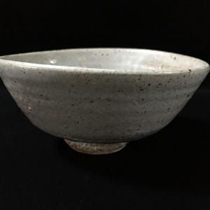 May include: A ceramic bowl with a speckled light gray glaze and a slightly irregular, organic rim. The exterior surface features subtle horizontal ridges and a visible, unglazed clay foot at the base, showcasing a natural, earthy texture.
