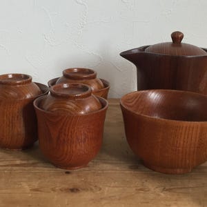 Op de afbeelding: Een houten theeservies bestaande uit een theepot met deksel, een open kom en drie kopjes met deksel. Elk stuk heeft een zichtbaar natuurlijk houtnerfpatroon in een middelbruine tint. De items zijn samen op een plat oppervlak geplaatst.