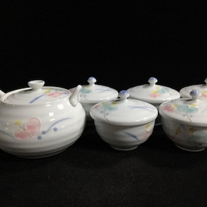 May include: A white ceramic tea set comprising one teapot and four lidded cups. Each piece features a delicate hand-painted floral motif in soft pastel shades of pink, blue, and yellow, with subtle horizontal ridges along the rounded bodies.