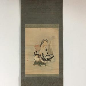 Op de afbeelding: Een verticale hangende rol met een schildering van een bebaarde figuur met een opvallend voorhoofd die een rode waaier vasthoudt. De figuur draagt donkere gewaden en een groene broek met patronen. Het kunstwerk is gemonteerd op donkergrijze stof.