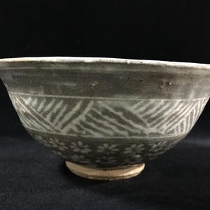 May include: A gray ceramic bowl with a white geometric pattern and small floral designs. The bowl has a slightly flared rim and a small, circular base. The design is repeated around the bowl's exterior.
