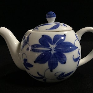 Op de afbeelding: Een witte keramische theepot met een handgeschilderd blauw bloemmotief en krullende ranken. De theepot heeft een rond lichaam, een gebogen tuit, een bijpassend deksel met een kleine blauwe knop en een gebogen handvat, versierd met blauwe blaadjes.