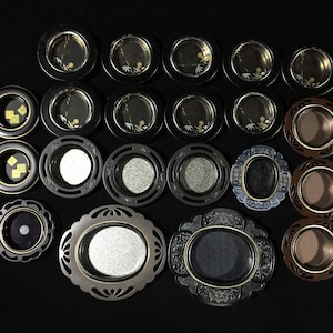May include: A collection of decorative, circular trays and dishes in various designs. Some have black or brown rims with gold accents, while others feature silver or gray finishes. The trays are arranged on a black surface.