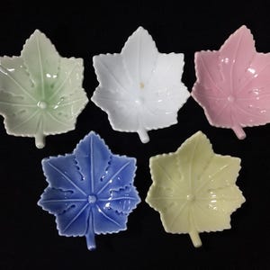 Könnte beinhalten: Ein Set aus fünf kleinen Keramikschalen in Form von Ahornblättern, die jeweils in einer anderen Pastellfarbe wie Hellgrün, Weiß, Rosa, Blau und Gelb gehalten sind. Jede Schale hat einen zentralen Stiel und geprägte Aderdetails.