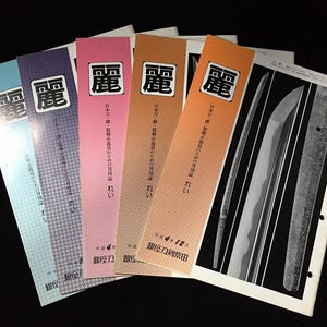 May include: A collection of Japanese magazines with colorful covers, featuring the kanji character "麗" in a white square. The magazines are in shades of blue, purple, pink, and orange. One magazine displays images of swords.