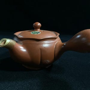 Könnte beinhalten: Eine braune Keramik-Teekanne mit einem langen, gebogenen Ausguss und einem abgerundeten Griff. Die Teekanne hat einen Deckel mit einem kleinen Knopf und einem dekorativen Etikett. Der Ausguss hat eine hellfarbige Spitze. Die Teekanne steht auf einer dunklen Oberfläche.