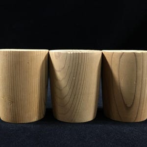 May include: Three cylindrical wooden cups with a natural wood grain finish. The cups are light brown and have a smooth, unadorned surface. The cups are of varying wood grain patterns and are set against a dark background.