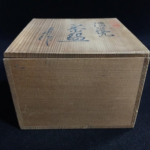 Op de afbeelding: Een rechthoekige houten opbergdoos met een lichtbruine houtnerfafwerking. Het deksel is voorzien van donkerblauwe Japanse kalligrafietekens in een verticale oriëntatie, samen met een kleine rode cirkelvormige stempel in de rechterbovenhoek.