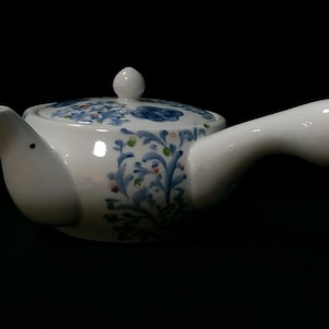 Könnte beinhalten: Weiße Keramik-Teekanne mit seitlichem Griff und Deckel. Die Teekanne ist mit blauen Blumenmustern und grünen und roten Akzenten verziert. Der Ausguss ist wie ein Vogelschnabel geformt.