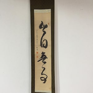 May include: A vertical hanging scroll featuring black ink calligraphy on a light beige paper background. The scroll is framed by a dark brown fabric border with a subtle pattern and includes red seal stamps and small black text along the side.