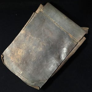 May include: A folded, weathered leather pouch in shades of gray and brown. The pouch appears to be handmade, with visible stitching along the edges. The leather has a worn, textured appearance, suggesting age and use. The background is black.