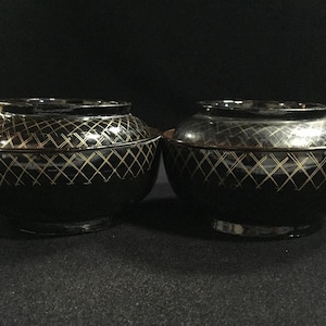May include: Two black lacquer bowls with lids. Each bowl has a gold crisscross pattern and a dark rim. The bowls are sitting on a dark surface. The lids are slightly domed and have a matching pattern.