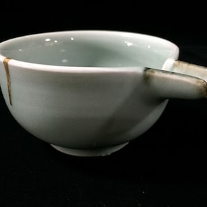 May include: A light blue ceramic pouring bowl with a smooth, glazed finish. The bowl features a distinct spout on one side and a thin, vertical gold-coloured line running down the exterior wall, indicating a repair along the side of the vessel.