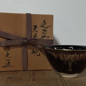 Puede incluir: Un cuenco de cerámica con esmalte marrón oscuro y patrones de goteo blancos cerca del borde, junto a una caja de almacenamiento de madera cuadrada atada con una cinta de tela marrón. La tapa de la caja muestra caligrafía japonesa en negro.