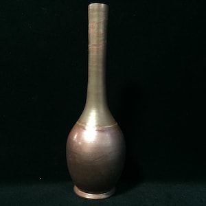 Pode incluir: Um vaso de cerâmica alto e esguio com um pescoço estreito e uma base arredondada e bulbosa. A superfície apresenta um acabamento metálico em tons de bronze escuro com estrias horizontais subtis e um brilho suave e reflexivo.