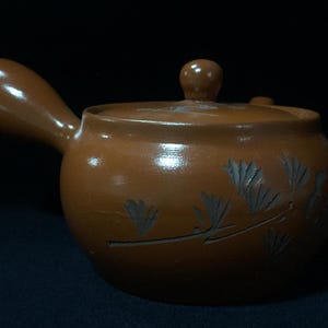 Könnte beinhalten: Eine braune Keramik-Teekanne mit Henkel und Deckel. Die Teekanne hat einen runden Körper und ist mit geätzten floralen Designs verziert. Der Griff ist gebogen und der Deckel hat einen kleinen Knopf. Die Teekanne steht vor einem dunklen Hintergrund.