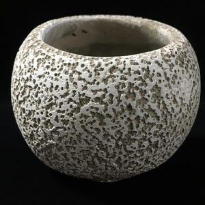 Puede incluir: Una maceta esférica con una textura de superficie rugosa, porosa y llena de cráteres. El exterior presenta un esquema de color de dos tonos con un acabado gris claro o blanco roto sobre una base de color marrón claro, creando un aspecto profundo.