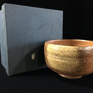 Op de afbeelding: Een ronde keramische kom met een textuurrijke, gevlekte bruine en beige glazuurafwerking en een licht taps toelopende voet. Naast de kom staat een rechthoekige, donkergrijze opbergdoos met een vezelige, papierachtige textuur en een klein wit label.