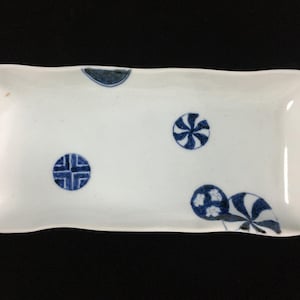 Puede incluir: Una bandeja de servicio rectangular de cerámica blanca con bordes ondulados, decorada con motivos circulares azules pintados a mano, incluyendo molinillos, patrones geométricos y diseños florales dispersos por la superficie.