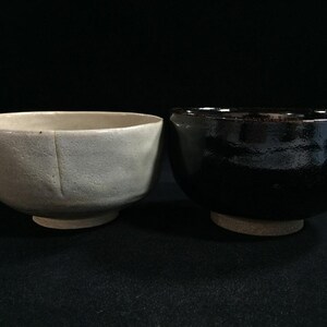 Op de afbeelding: Twee keramische kommen. Eén is lichtbeige van kleur met een textuuroppervlak en een zichtbare naad. De andere kom is zwart met een glanzende afwerking. Beide kommen hebben een afgeronde vorm en staan op een kleine, cirkelvormige basis.
