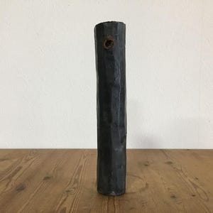 May include: A tall, cylindrical, dark charcoal-gray vase with a textured, uneven surface. The vase features a single, small, circular opening near the top rim, showcasing a weathered, rusted-brown metallic finish around the hole.