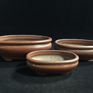 May include: Three oval-shaped bonsai pots in varying sizes, crafted from unglazed reddish-brown clay. Each pot features a smooth, rounded rim, a shallow basin, and small, integrated feet at the base, presenting a consistent earthy tone throughout.