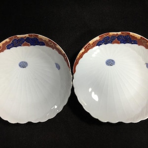 Puede incluir: Dos platos de cerámica blanca con borde festoneado. Cada plato presenta un diseño radial que emana de un punto azul central. Los bordes están decorados con un patrón floral marrón y azul. Los platos se muestran sobre una superficie negra.
