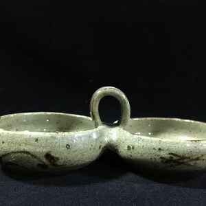 May include: A ceramic serving dish with two connected bowls and a handle. The dish is a muted green colour with dark brown accents. The handle is a small, circular loop.