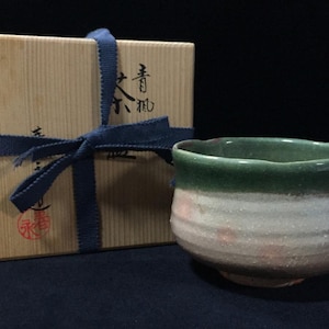 Op de afbeelding: Een keramische theekom met een groen geglazuurde rand en een textuurrijke witte en beige kom, vergezeld van een vierkante houten opbergdoos vastgebonden met een blauw stoffen lint. De doos bevat zwarte Japanse kalligrafie en een rond rood stempel.
