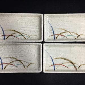 May include: A set of four rectangular ceramic plates with a speckled off-white glaze. Each plate features a hand-painted design of thin, curved lines in blue, brown, red, and teal, resembling blades of grass or reeds against a light background.