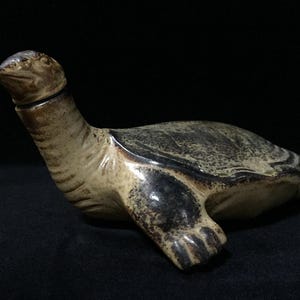 Op de afbeelding: Een keramisch schildpadvormig vat met een afneembaar deksel in de vorm van een kop. Het object heeft een gespikkeld glazuur in tinten donkerbruin, antraciet en crème, met een getextureerd, verweerd uiterlijk op het schild en de ledematen.