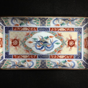 Könnte beinhalten: Ein rechteckiges Keramiktablett mit einem zentralen Medaillon, das einen stilisierten Vogel zeigt, umgeben von komplizierten Blumenmustern und geometrischen Rändern in Blau, Rot, Grün und Gold. Der Rand ist mit einer dünnen Goldkante versehen.
