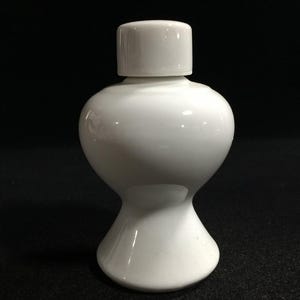 Puede incluir: Un tarro de cerámica blanco brillante con un cuerpo redondeado y bulboso y una base estrecha y acampanada. El recipiente presenta una superficie lisa y reflectante y está rematado con una tapa cilíndrica a juego que encaja en la abertura.