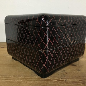 Pode incluir: Uma caixa quadrada de dois níveis em laca preta com um padrão de treliça vermelho repetitivo nas superfícies externas. O recipiente tem cantos arredondados, um acabamento liso e brilhante e uma costura distinta que separa as duas seções.