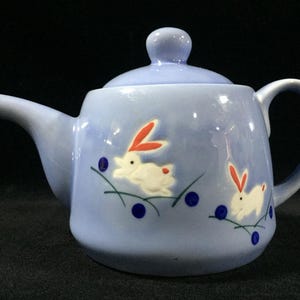 May include: A light blue ceramic teapot with a lid and handle. The teapot is decorated with white rabbits with red ears and blue dots. The teapot is designed for serving tea.