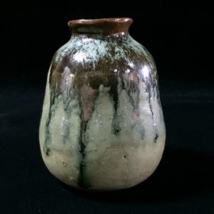 Peut inclure: Un petit vase en céramique avec un corps arrondi et un col étroit. La surface présente une glaçure réactive avec un sommet brun foncé qui coule sur une base vert clair texturée, créant un aspect tacheté et superposé sur l'ensemble.