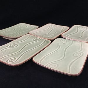 May include: A set of five rectangular ceramic plates featuring a light sage green glaze with a raised, embossed pattern of swirling lines and gentle waves. Each plate has a smooth, slightly curved surface and a distinct dark brown rim.