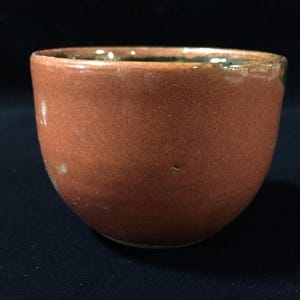 May include: A small, round ceramic bowl with a smooth, matte, reddish-brown exterior finish. The rim of the bowl shows a subtle, dark, glossy glaze that transitions into the interior, which appears dark and reflective against the solid dark background.