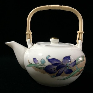 Puede incluir: Una tetera de porcelana blanca con un diseño pintado de flores de iris moradas, hojas verdes y detalles dorados. La tetera presenta un pico curvo, una tapa a juego con un pomo dorado y un asa móvil de color claro con textura estriada.