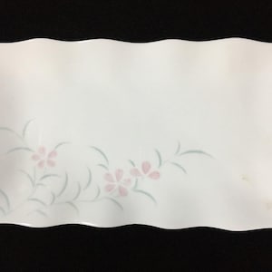 May include: A rectangular white ceramic serving platter with wavy, scalloped edges. The surface features a delicate, muted illustration of light pink flowers with soft green stems and leaves positioned in the lower left corner.