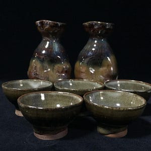 以下が含まれることがあります： 陶器製の徳利2本と、お揃いの小さな猪口5個のセット。徳利は茶色、青、黄褐色の光沢のあるまだら模様の釉薬が特徴です。猪口は、土のような緑と茶色の斑点模様の仕上げで、内側は滑らかな釉薬が施されています。