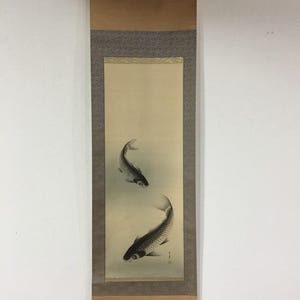 May include: A traditional Japanese scroll painting featuring two koi fish in shades of gray and black. The artwork is set against a light beige background, framed by a patterned border and brown paper.