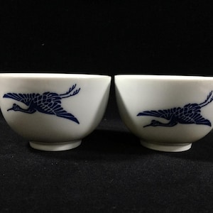 May include: Two white ceramic tea cups with a blue crane design. The crane is depicted in flight, with its wings outstretched. The cups have a rounded shape and a small base. The background is black.