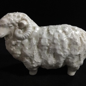 May include: A white ceramic figurine of a ram with a glossy, textured finish that mimics the appearance of thick, curly wool. The sculpture features curved horns on the side of the head and stands on four short legs, captured in a side profile view.
