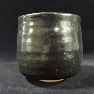 Peut inclure: Une tasse en céramique faite à la main, à glaçure foncée, avec un corps arrondi et un bord légèrement évasé. La tasse présente une surface texturée avec de subtiles variations de couleur, allant du noir profond à des nuances de brun et de gris. La base est non émaillée.