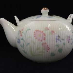 Puede incluir: Una tetera de porcelana blanca con una delicada textura acanalada y un patrón floral pintado en tonos suaves de rosa, azul y verde. La tetera incluye una tapa a juego con un pomo de tono dorado y un pico curvo en el lado izquierdo.