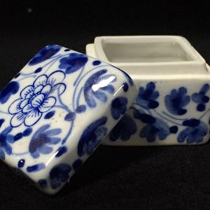 Pode incluir: Uma pequena caixa quadrada de cerâmica com tampa removível, apresentando uma base branca decorada com padrões florais azuis pintados à mão. A tampa está posicionada num ângulo ao lado do recipiente aberto, exibindo pétalas azuis detalhadas.